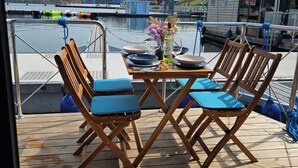 Eine Holzterrasse mit zwei Klappstühlen und einem Tisch, mit Blick auf ein Gewässer mit Booten und Gebäuden im Hintergrund.