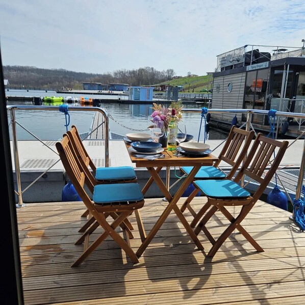A wooden deck with two folding chairs and a table, overlooking a body of water with boats and buildings in the background.