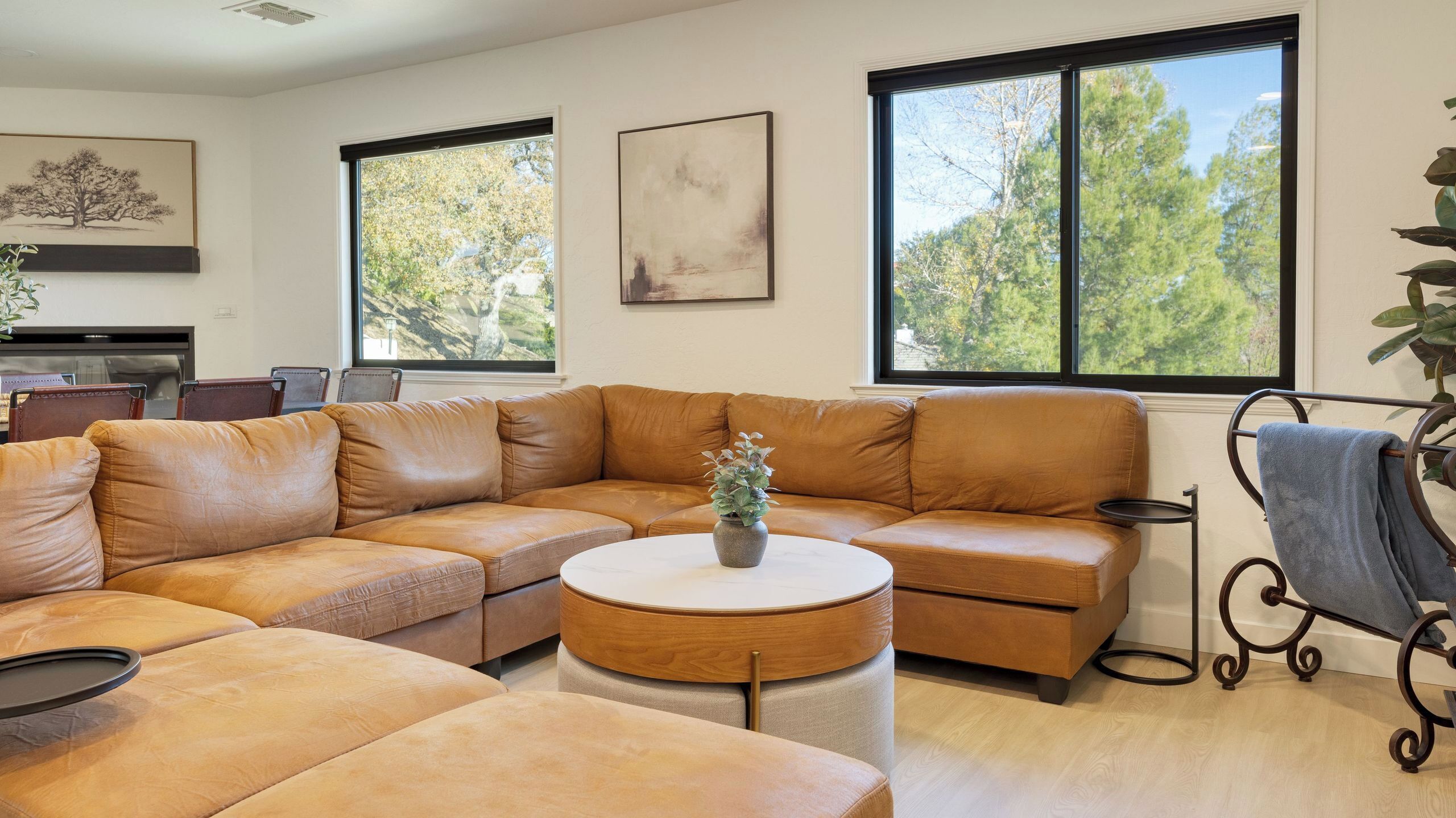 Ein modernes Wohnzimmer mit einer großzügigen L-förmigen Couch, einem runden Couchtisch und Blick auf Bäume durch große Fenster.