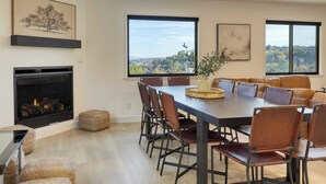 Ein Essbereich mit großem Tisch, Stühlen, einem Kamin und großen Fenstern mit Blick ins Freie.