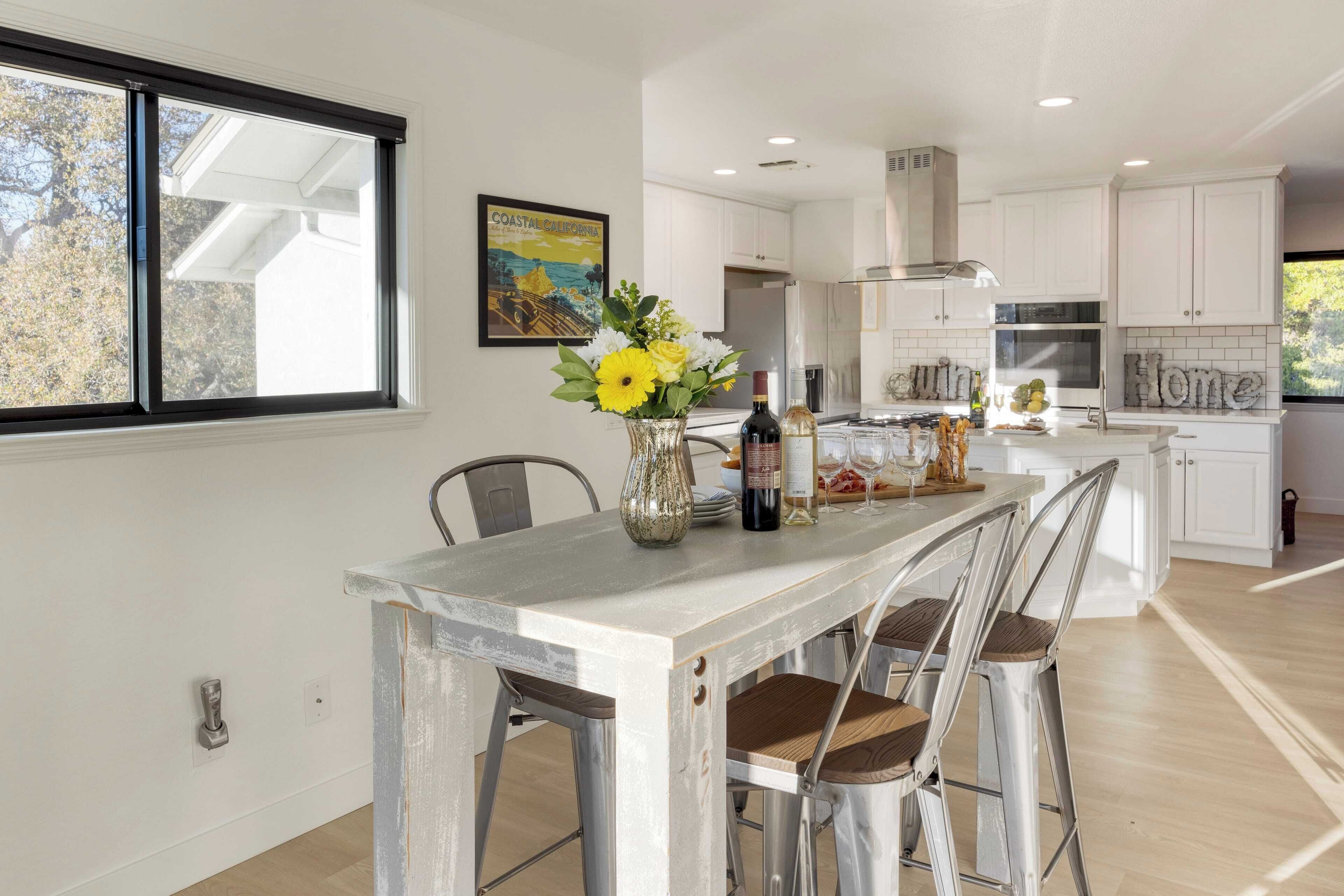 A modern kitchen with white cabinets, a central island with bar stools, a window with a view of trees, and a framed picture on the wall.