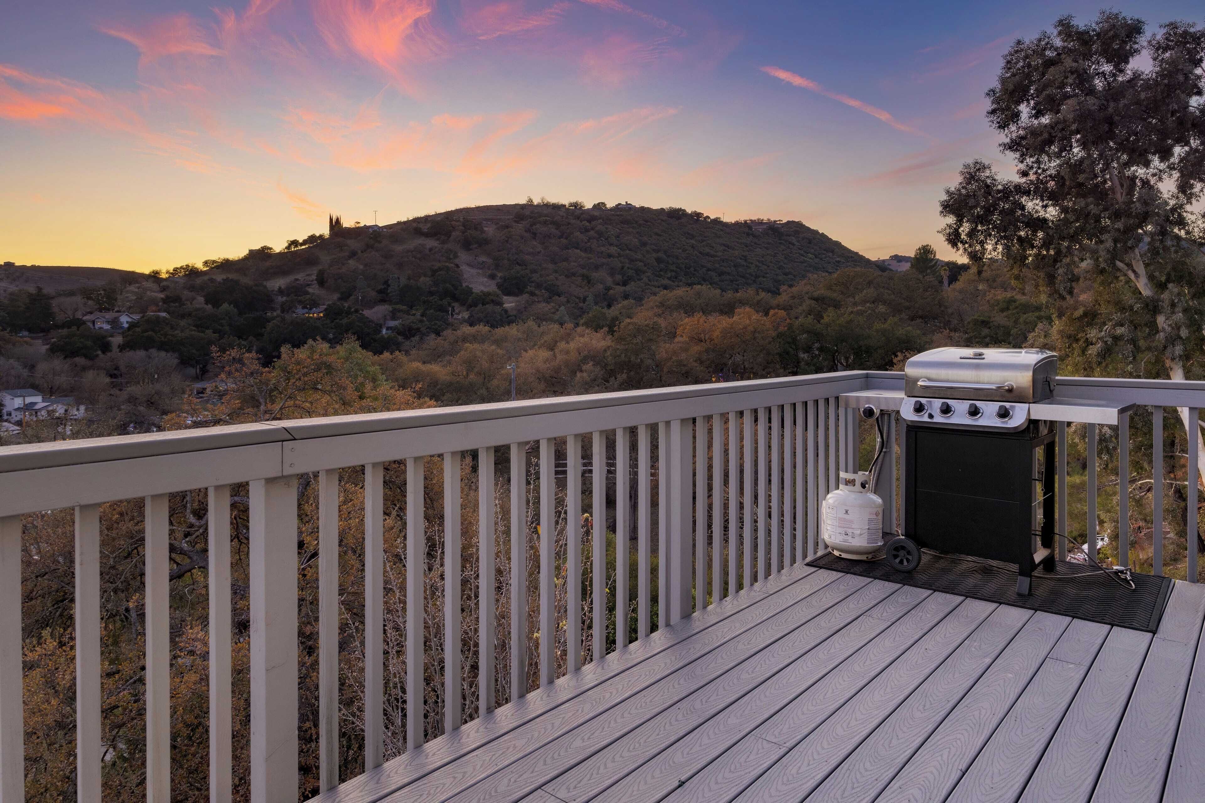 A deck with a barbecue grill and propane tank, overlooking a hilly landscape with trees and a distant hill.