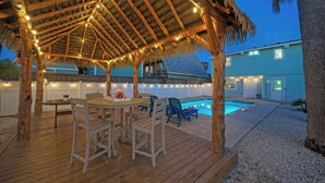 Une terrasse en bois surmontée d’un toit de chaume, avec des sièges d’extérieur et une piscine.