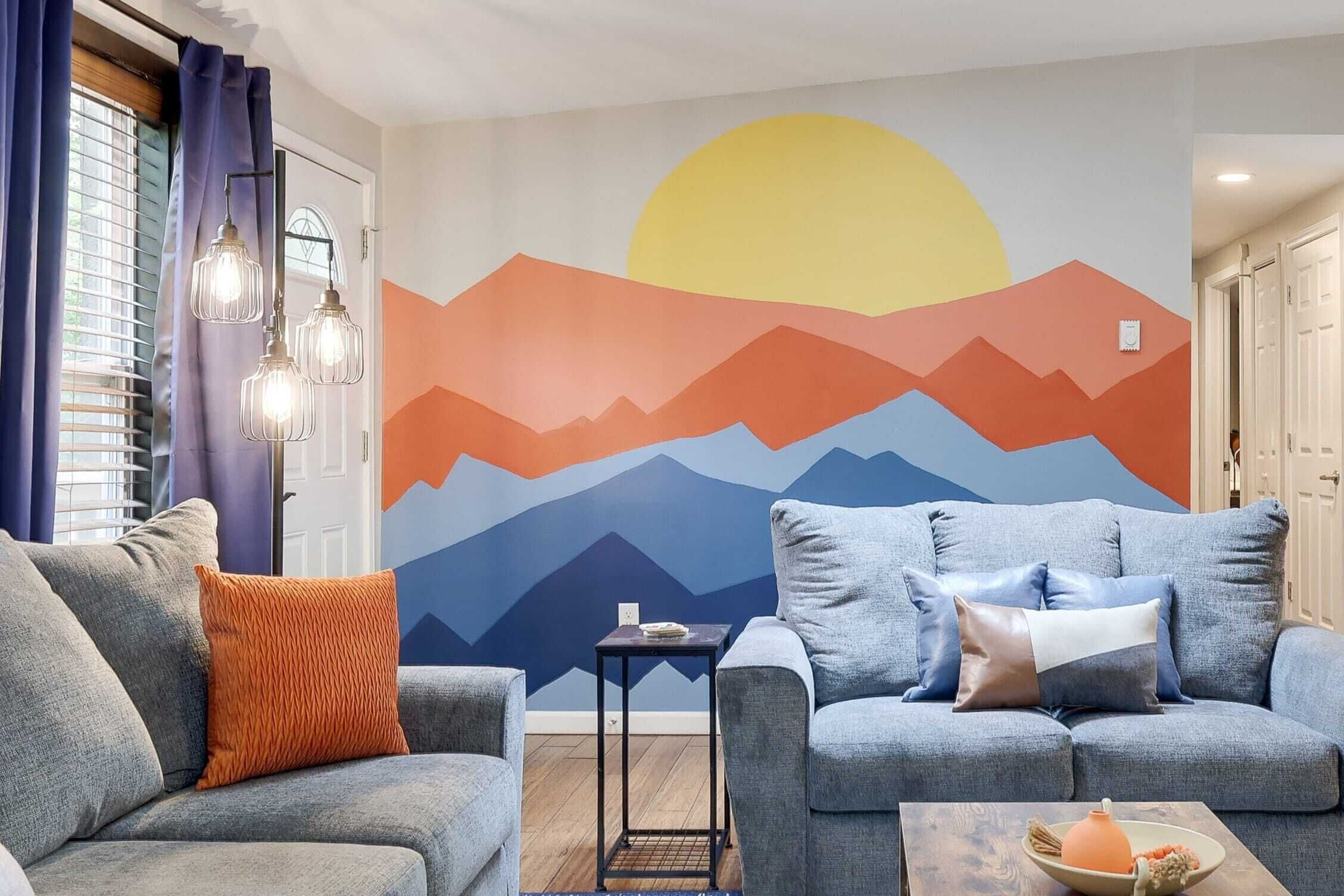 A living room with two grey sofas, one with an orange cushion, and a small table with a bowl and a teapot. A wall mural depicts a sunset over mountains.