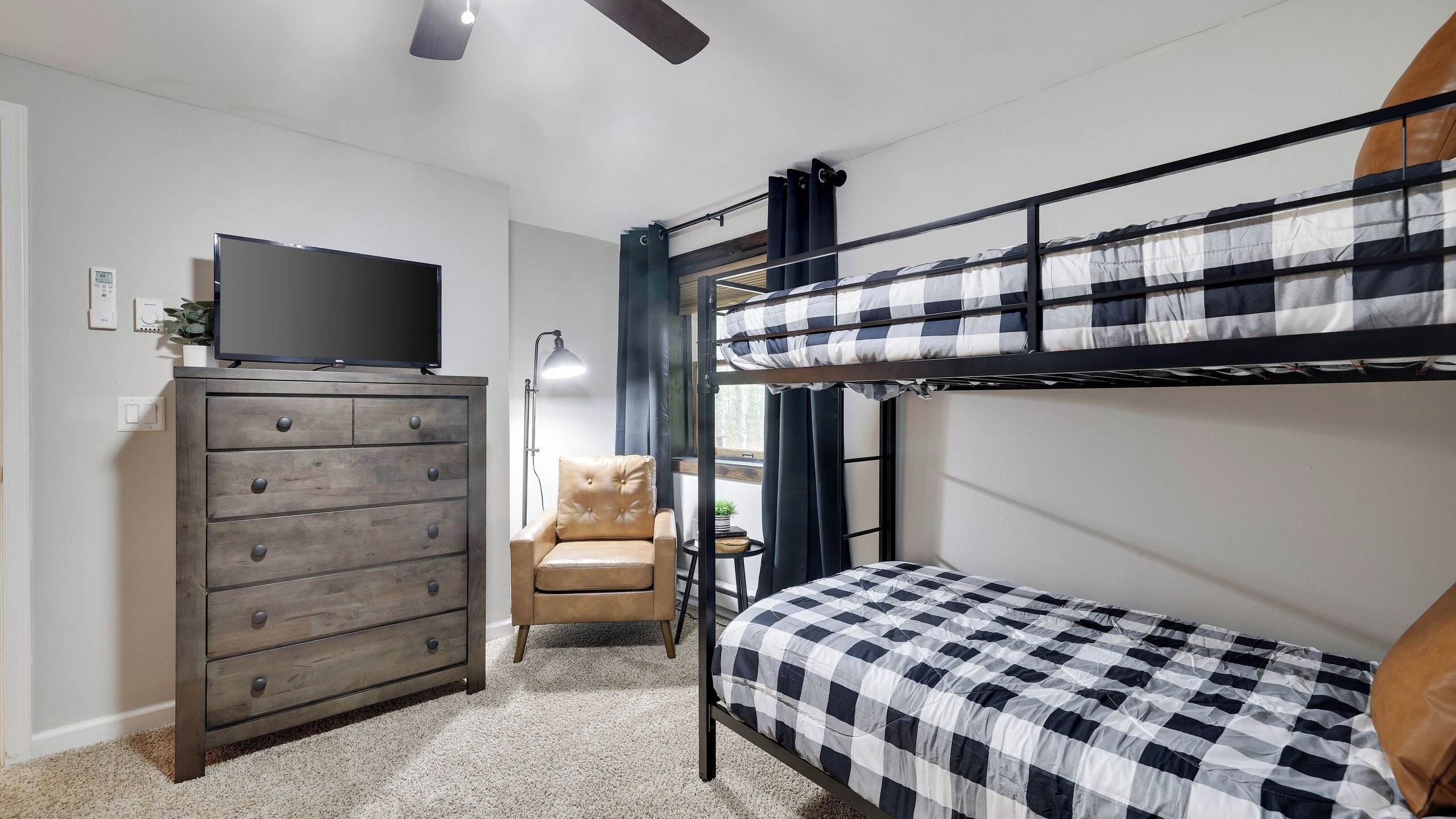 A bunk bed with a checkered bedspread, a dresser with drawers, a television on top, a chair, and a small table with a lamp.