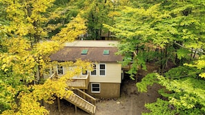 A small, single-story house surrounded by dense trees with a wooden deck and a small staircase leading to the entrance.