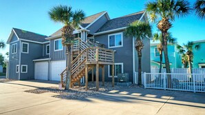 A two-story house with a wooden staircase leading to a deck, surrounded by palm trees and a white fence. - Communal Pool, Beach Gear & Prime Location Near Port Aransas Beaches Salty Seas by Avantstay (Port Aransas)