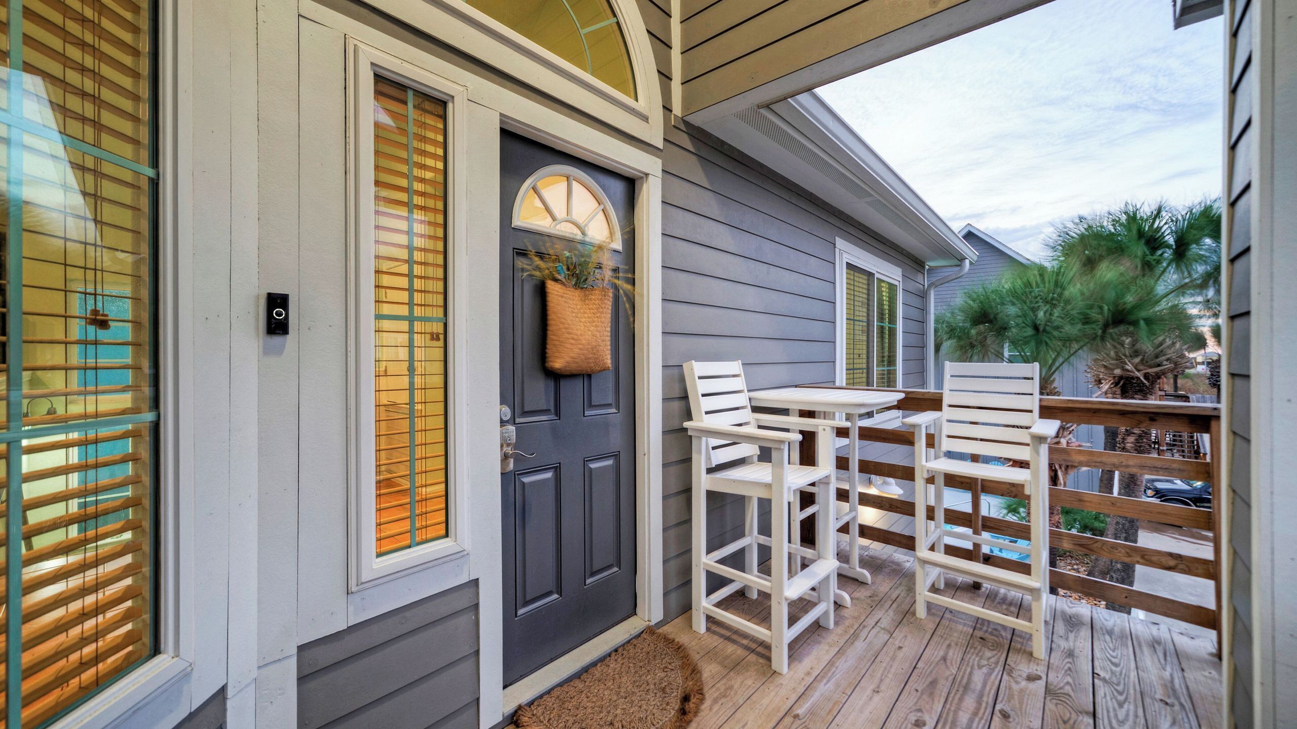 A wooden deck with a table and chairs, a dark door with a basket, and a window with blinds.