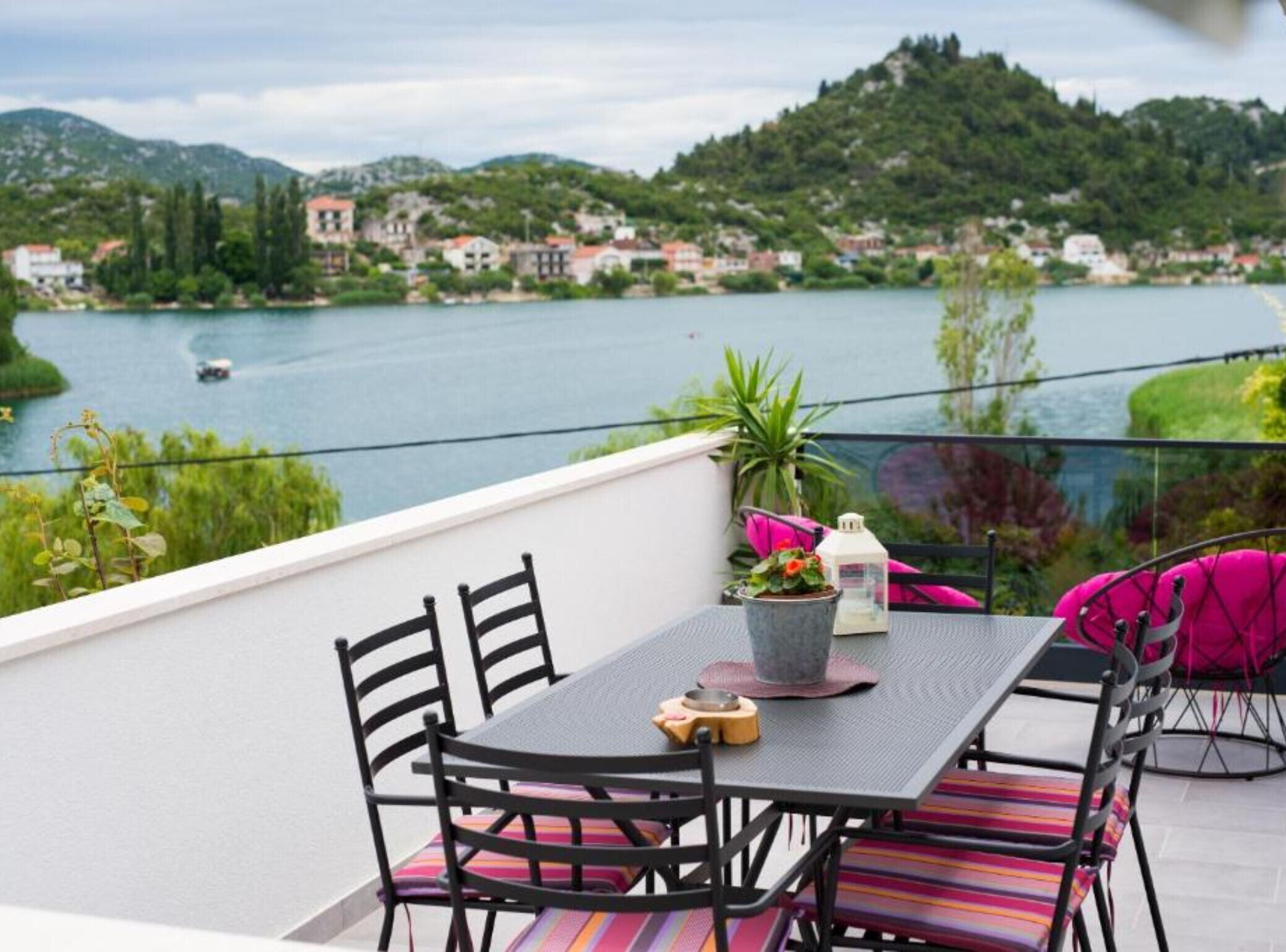 A balcony with a table and chairs overlooking a lake, surrounded by greenery and buildings.