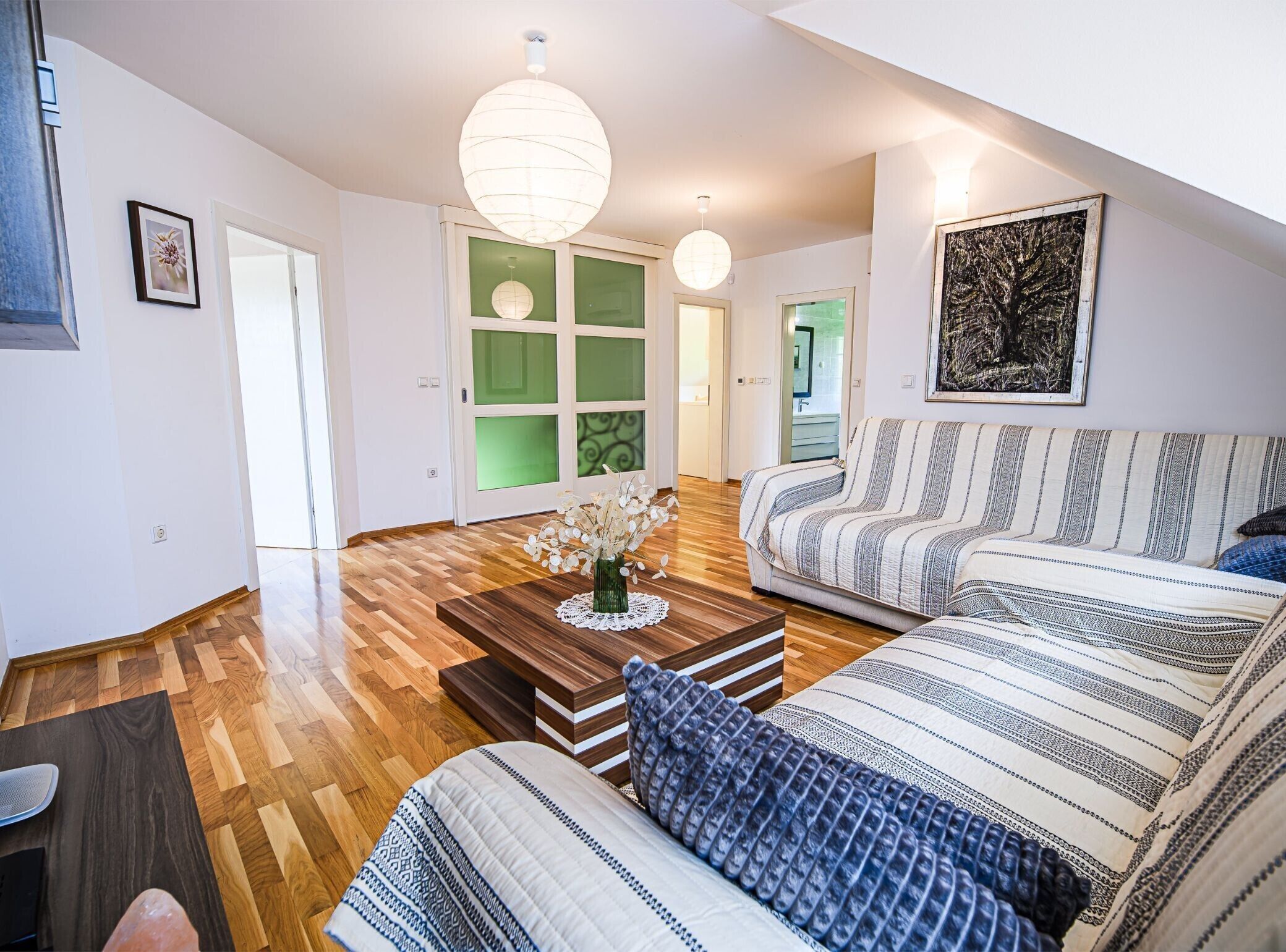 A modern living room with a wooden parquet floor, a striped sofa, a coffee table with a vase, and a green-framed door.