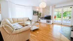 A modern living room with a beige sofa, a white coffee table, a TV on a white stand, and a large window with a view of greenery. - Stunning Rural Holiday Home with Garden and BBQ - Four Bedroom Holiday Home (Brežice)