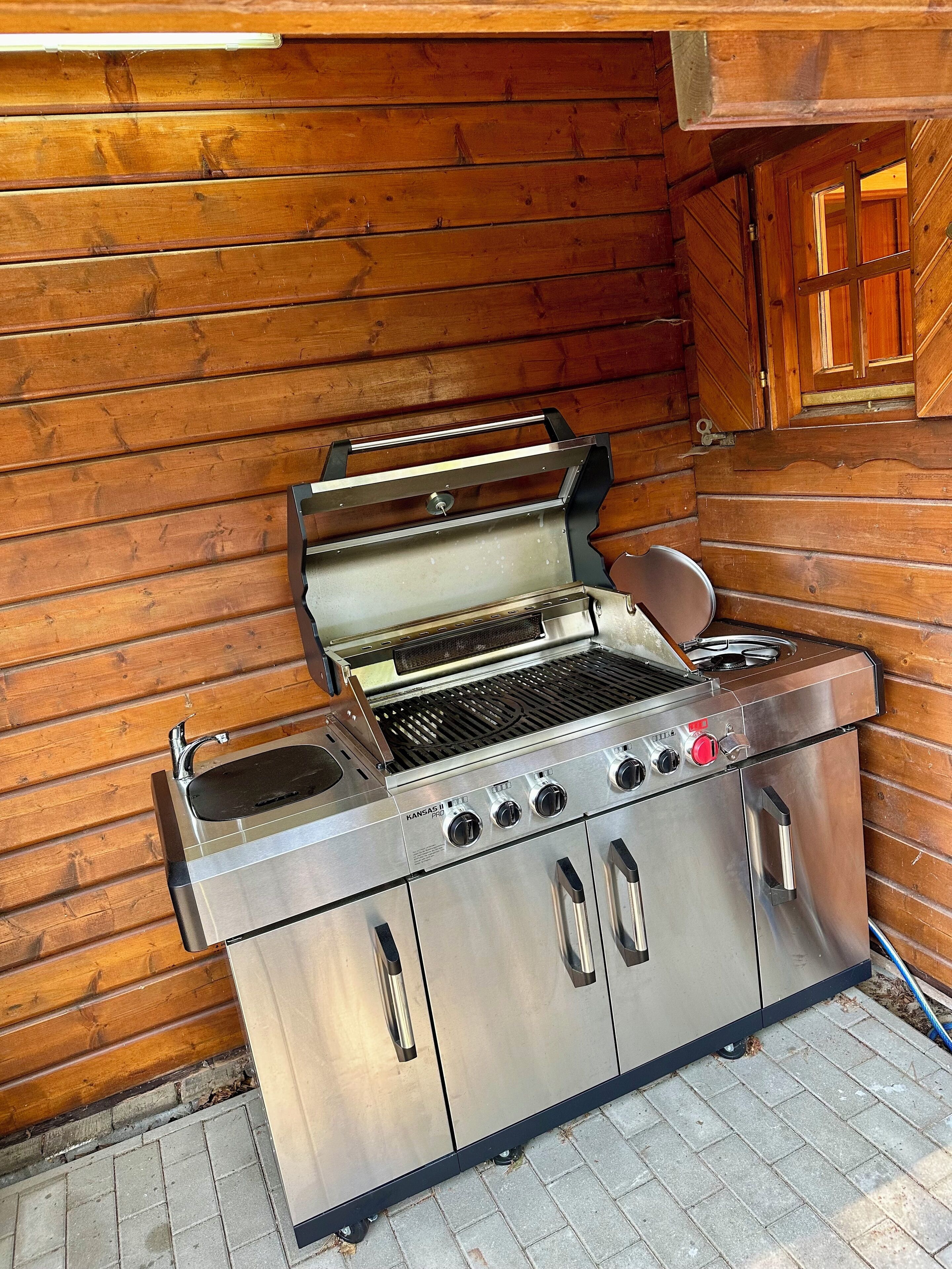 A stainless steel barbecue grill with a sink and a cabinet, set against a wooden wall.