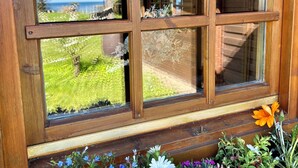 A wooden window with multiple panes, a flower box with various flowers, and a view of a wooden building and greenery. - Almhuetten-am-Meer.de (Lubmin)