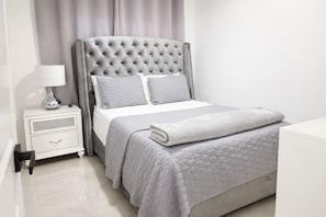 A neatly arranged bedroom with a tufted headboard, a bedside table with a lamp, and a bed with a quilted blanket.
