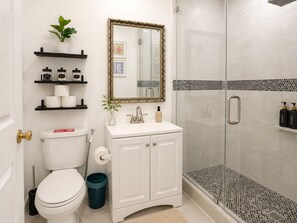 A bathroom with a toilet, sink, mirror, shelves with toiletries, and a shower area.
