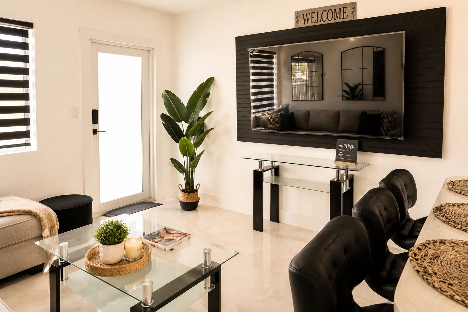 Una sala de estar moderna con una mesa de centro de vidrio, un sofá, un sillón de cuero negro, una planta en maceta, un televisor y un letrero que dice 'WELCOME'.