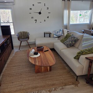 Uma sala de estar com mesa de centro de madeira, sofá, relógio na parede e janela com cortinas.