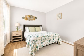 Quarto com cama, mesa de cabeceira, luminária, arte decorativa na parede e um tapete pequeno.