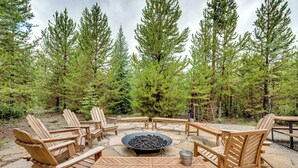 A circular seating area with a central fire pit, surrounded by wooden Adirondack chairs and tables, set on a stone patio with a forest backdrop. - Mtn Escape w/ Hot Tub, By Trails in Grand Lake! (Grand Lake)