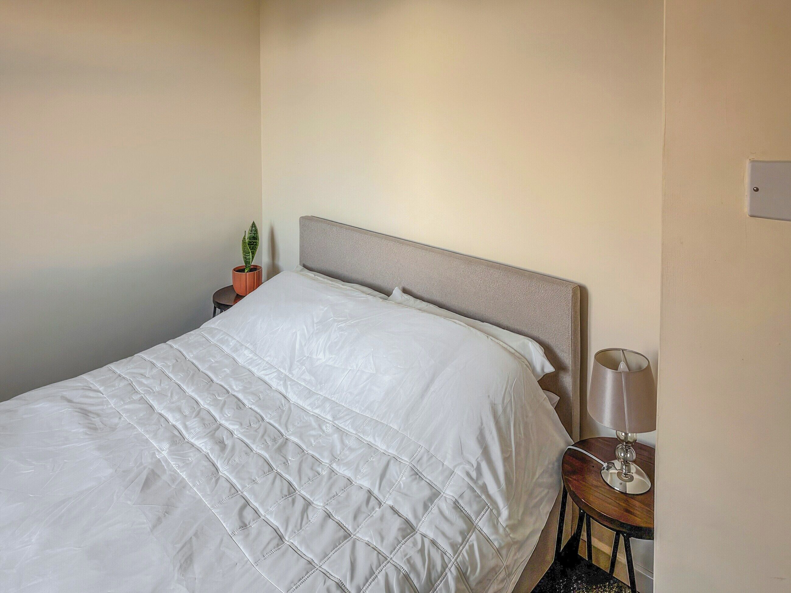 Ein ordentlich bezogenes Bett mit weißer, gesteppter Daunendecke, einem grauen Kopfteil, einem kleinen Topfpflanzen und einem Nachttisch mit einer Lampe.