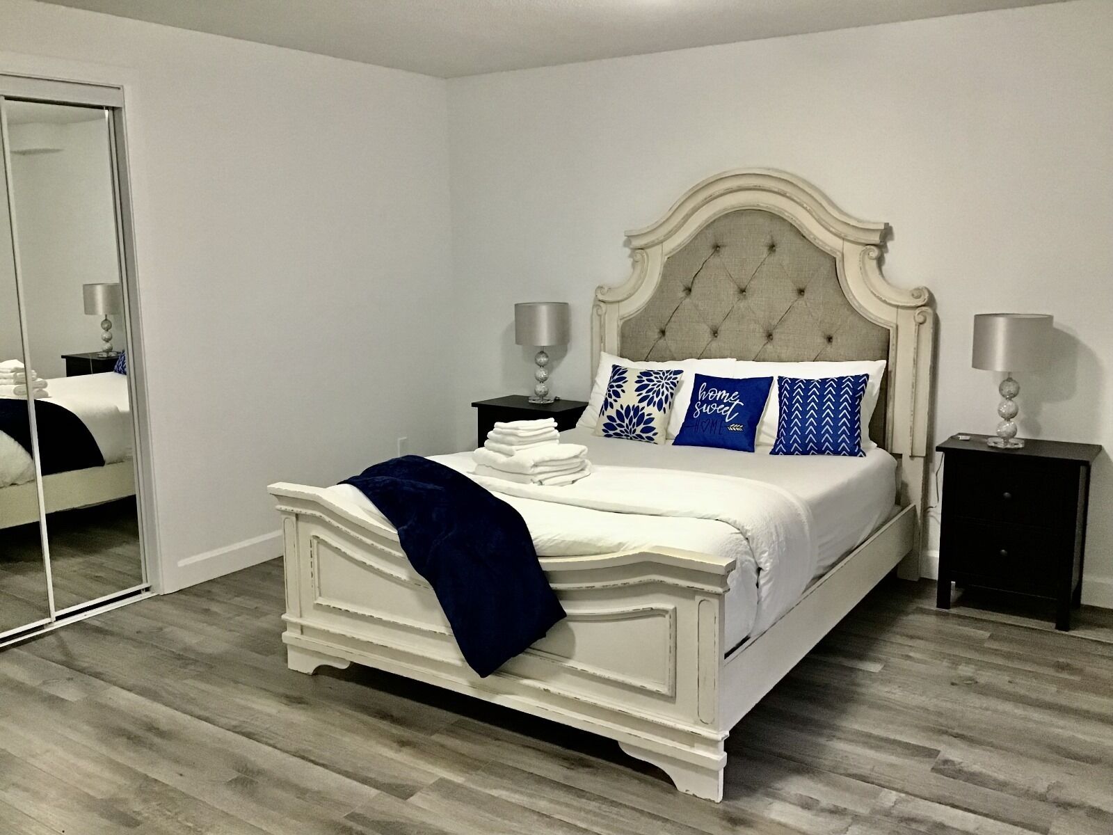 Une chambre à coucher avec un lit blanc, un plaid bleu foncé et des coussins décoratifs. Il y a deux tables de chevet avec des lampes.