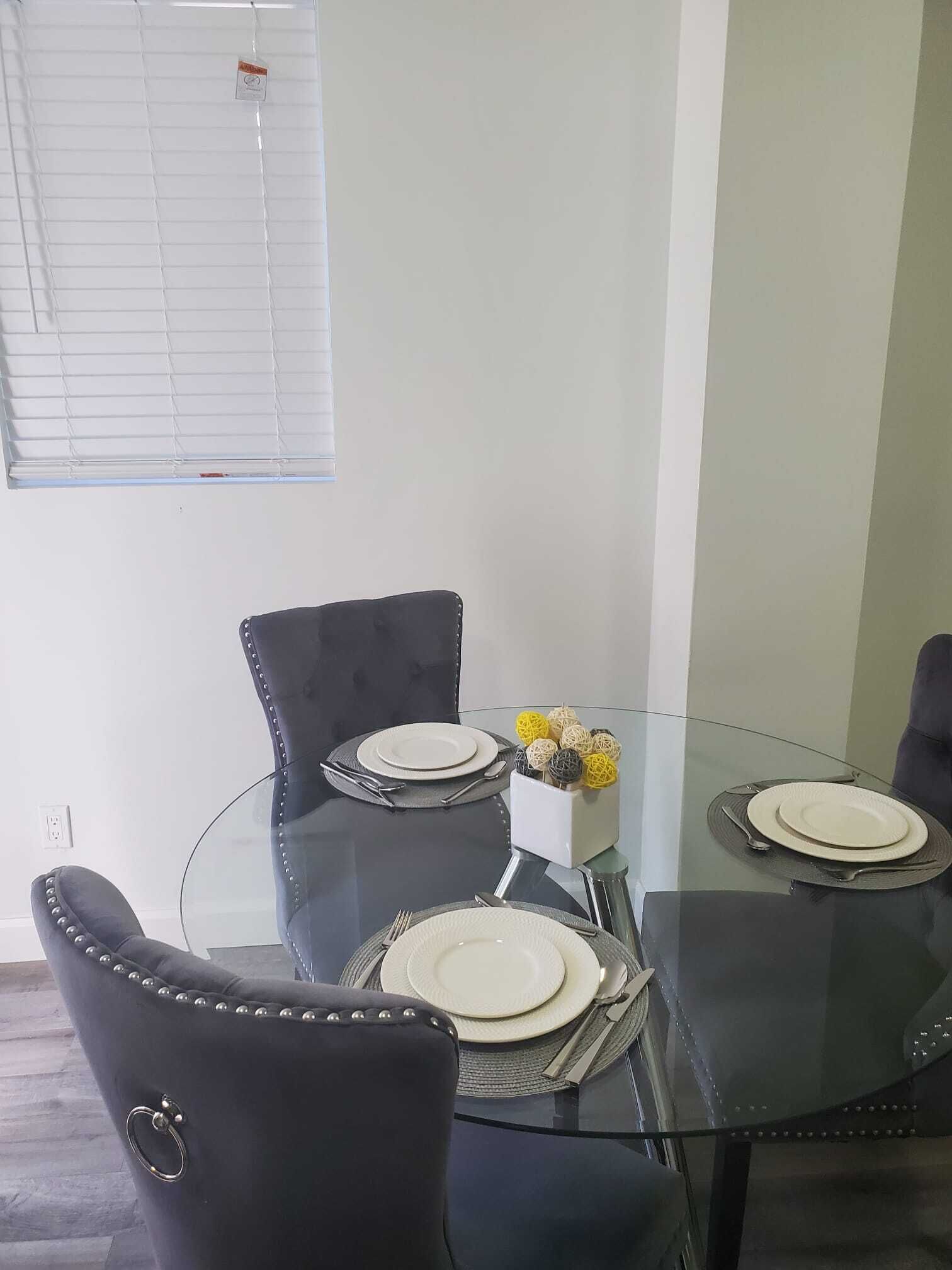 A dining area with a glass table surrounded by four black chairs with metallic studs. There are plates and cutlery on the table, and a small decorative item in a white box.