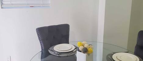 A dining area with a glass table surrounded by four black chairs with metallic studs. There are plates and cutlery on the table, and a small decorative item in a white box.