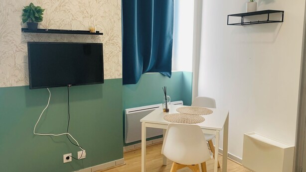 A room with a flat-screen TV mounted on the wall, a white table with two chairs, a small shelf with a plant, and a blue curtain. - T1 bis for 3 people Private and separate kitchen bathroom (Saint-Brieuc)