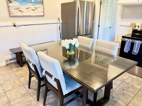 A modern kitchen with a glass-top dining table, four upholstered chairs, a stainless steel refrigerator, and a black oven.