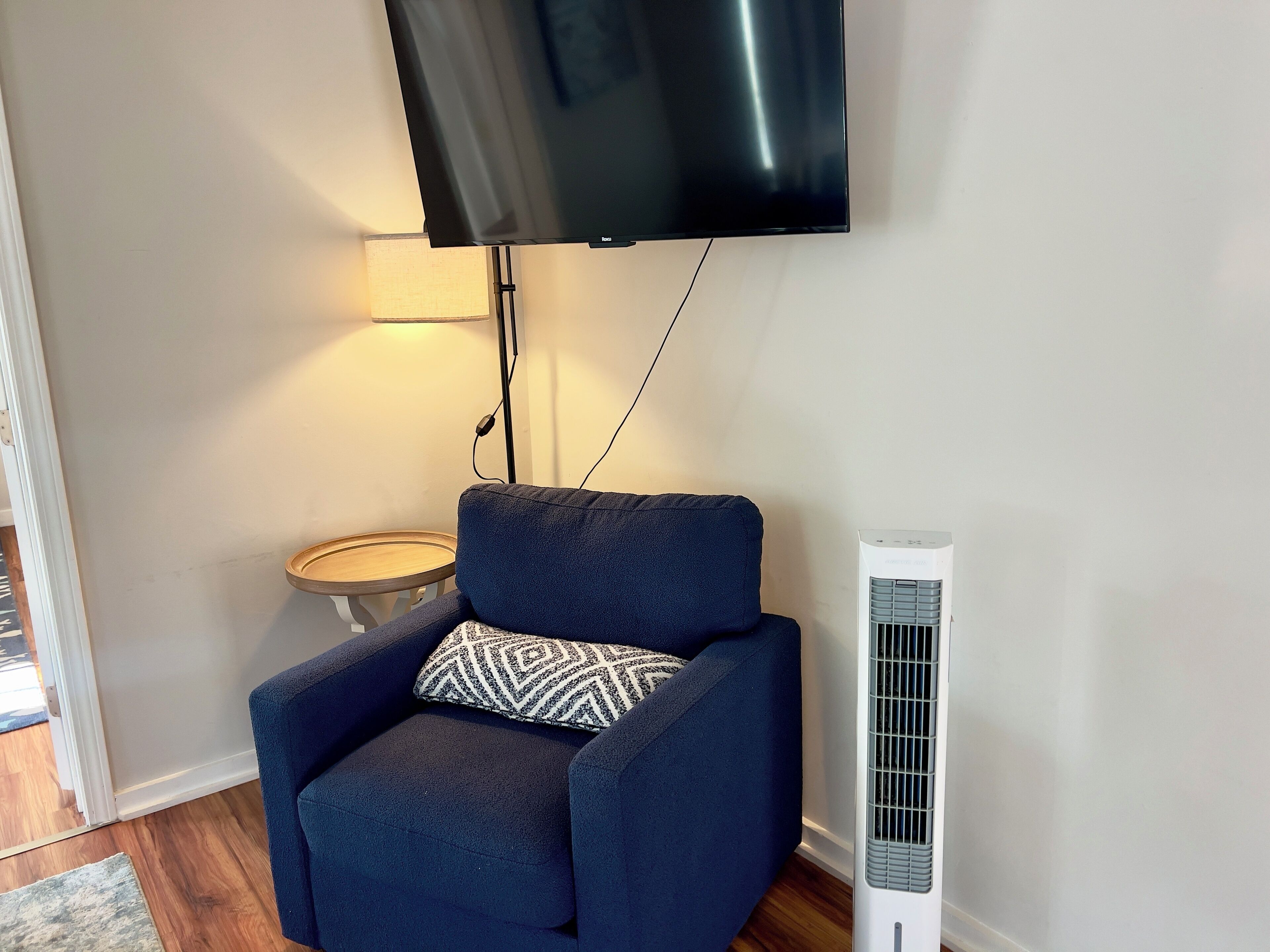 A living room with a dark blue armchair, a patterned pillow, a standing heater, a small round table with a lamp, and a flat-screen TV mounted on the wall.
