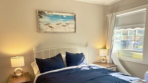 A bedroom with a bed, two bedside tables, a nightstand, a window with curtains, and a painting on the wall. - Relax and unwind in our 4 bedroom beach condo. (Old Orchard Beach)