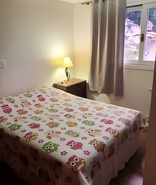 Un dormitorio con una cama cubierta con una colcha estampada de animales de colores, una lámpara de noche, una mesita de noche y una ventana con cortinas.