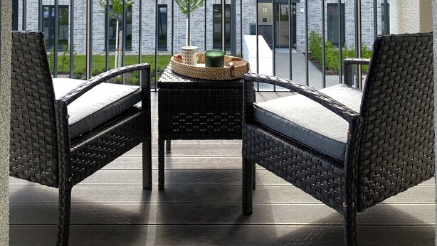 A balcony with wicker furniture, a small table, and a view of a modern building with multiple windows. - Luxurious 2-room apartment, WLAN, washing machine (Schönefeld)