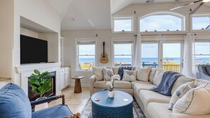 A living room with a fireplace, a television, a guitar, a sofa, and large windows overlooking the ocean. - Sleeps 28 Beachfront w/ Pool, Hot Tub, Game Room Atlantic Watch by Avantstay (Kill Devil Hills)