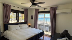 A bedroom with a bed, a ceiling fan, a window with curtains, a nightstand, and a view of the beach.