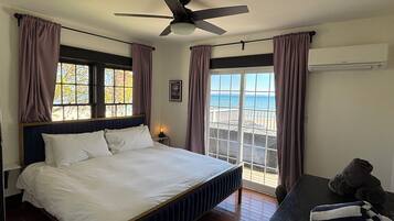 A bedroom with a bed, a ceiling fan, a window with curtains, a nightstand, and a view of the beach.