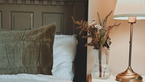 Een net opgemaakt bed met een groen kussen, een nachtkastje met een lamp en een vaas gedroogde bloemen, en een houten nachtkastje met lades.