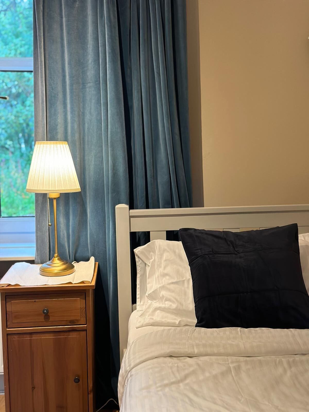 Ein ordentlich bezogenes Bett mit weißen und schwarzen Kissen, ein Nachttisch mit Lampe und ein Fenster mit blauen Vorhängen.
