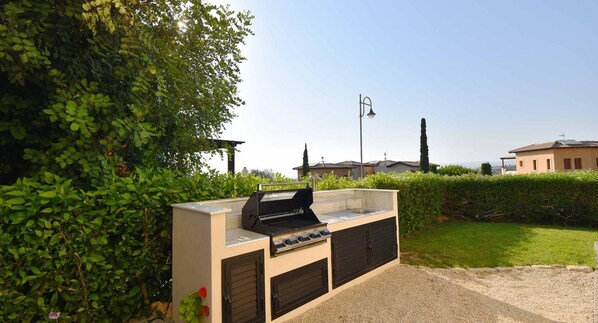 An outdoor kitchen with a grill, sink, and storage cabinets, surrounded by greenery and a clear sky.
