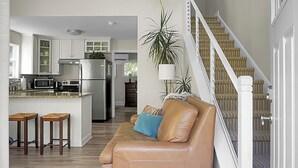 A spacious living area with a leather sofa, wooden stools, a kitchen with white cabinets, and a staircase with striped carpet. - Charming cottage! Central to beach & downtown (Mount Pleasant)