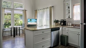 A kitchen with a view of the outdoors, a dining area with chairs, and a TV displaying a beach scene. - Charming cottage! Central to beach & downtown (Mount Pleasant)