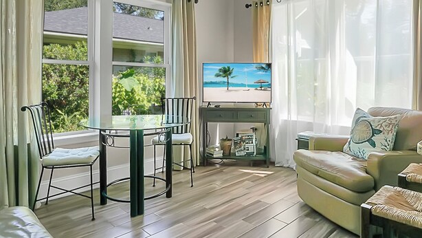 A living room with a glass table, two chairs, a sofa, a TV, and a view of greenery outside. - Charming cottage! Central to beach & downtown (Mount Pleasant)