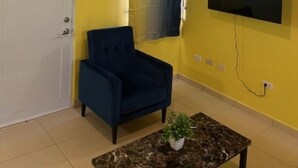 Ein Zimmer mit gelben Wänden, einem Deckenventilator, einem an der Wand montierten Fernseher, einem dunkelblauen Sessel, einer geschlossenen weißen Tür und einem kleinen Tisch mit einer Pflanze.