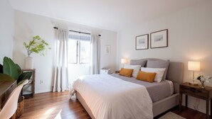 Un dormitorio con una cama grande, lámparas de noche, una mesita con lámpara, una planta y una ventana con cortinas.