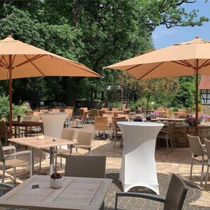 An outdoor patio area with tables and chairs, large umbrellas, and lush greenery in the background. - Hotel Le Filou  (Hille)