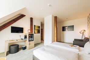 A modern bedroom with a loft-style ceiling, a large bed, a desk with a computer, and a TV mounted on the wall. - Hotel Le Filou  (Hille)
