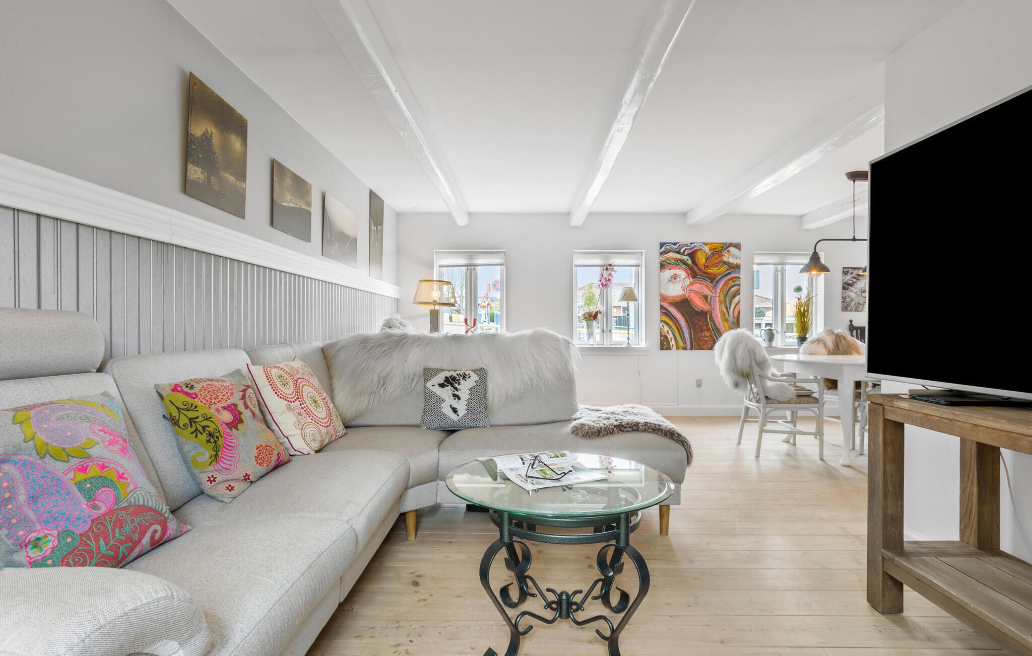 A modern living room with a grey sofa, colorful cushions, a glass coffee table, and a TV on a wooden stand.
