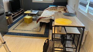 Une chambre à coucher de taille modeste, comprenant un lit, des tables de chevet, un téléviseur et une fenêtre donnant sur des immeubles.