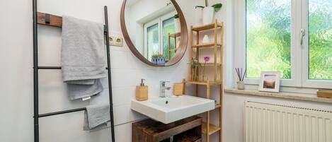 A bathroom with a wooden towel rack, a round mirror, a white sink, and a wooden shelf with a potted plant.