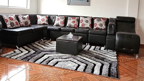Ein Wohnzimmer mit einem schwarzen Ecksofa, dekorativen Kissen mit Muster, einem schwarzen Couchtisch und einem Teppich mit Muster. Der Boden ist mit einem geometrischen Fliesenmuster versehen.